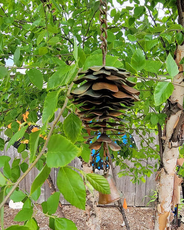 A metal pine cone wind chime hanging from a tree branch in a backyard. The feeder features a stacked, tiered design resembling pine cones in a weathered bronze-brown finish, with a small hanging metal bell below.