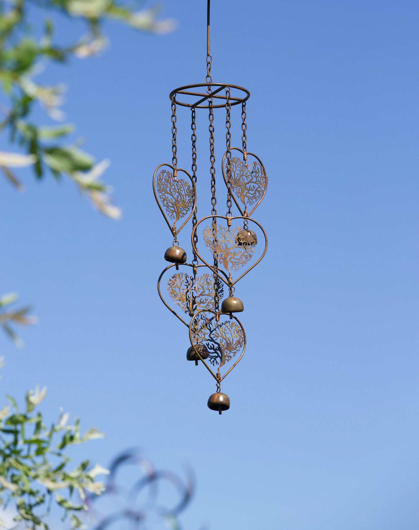 Antique bronze metal wind chime with heart-shaped tree of life cutouts and small bells hanging on chains against blue sky