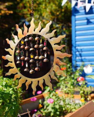 A sun-shaped metal wind chime hanging in a garden. The center features a circular black section filled with small hanging bells in various colors, surrounded by a gold-toned sunburst frame.