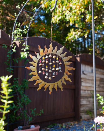A sun-shaped metal wind chime hanging from a garden arch in a backyard. The spinner features a gold-toned sunburst frame, a dark center, and multiple small hanging beads in various colors.