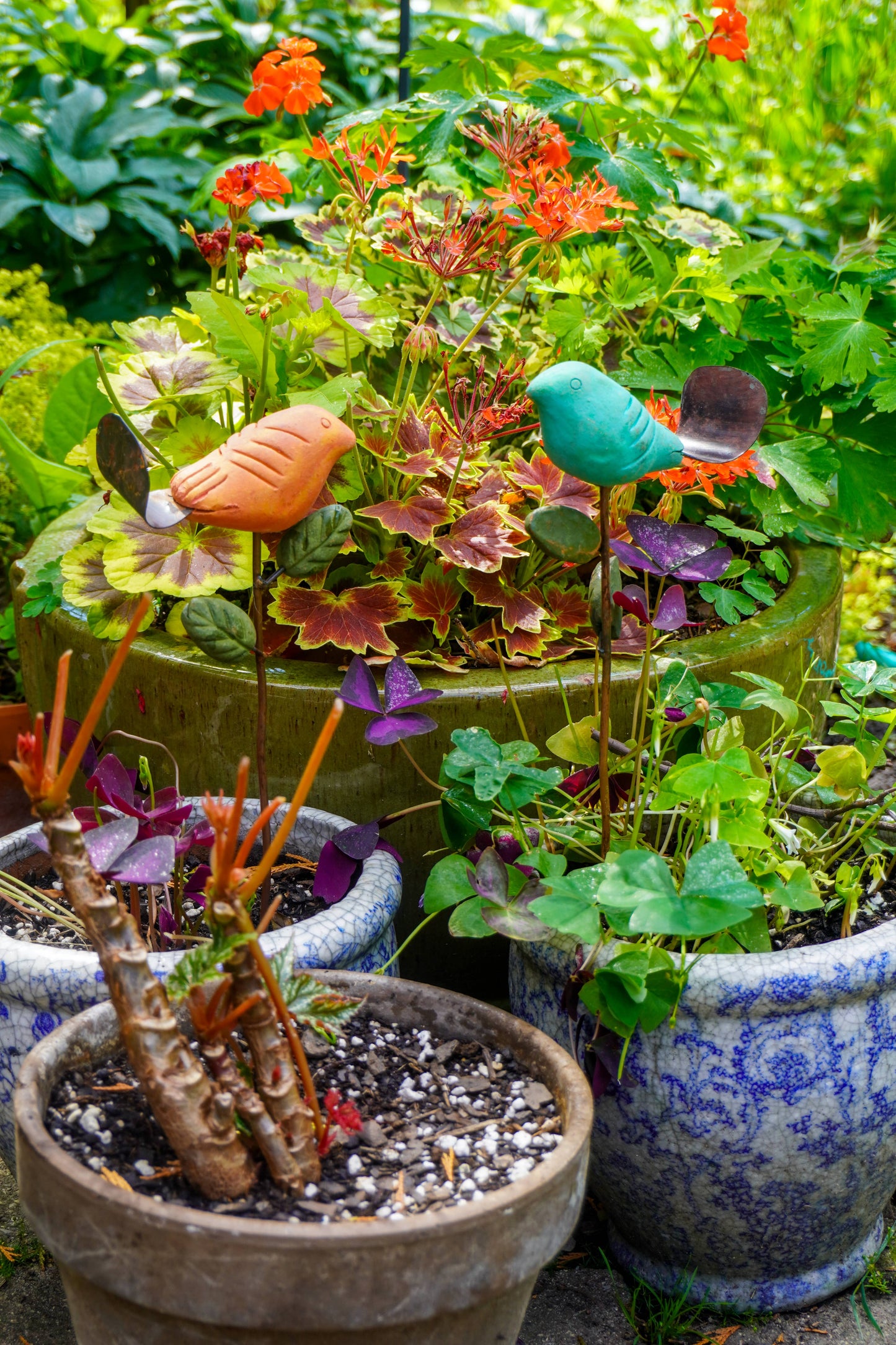 Rusty orange terracotta bird figurine on a metal stake with green cast leaf accents, placed among fern plants