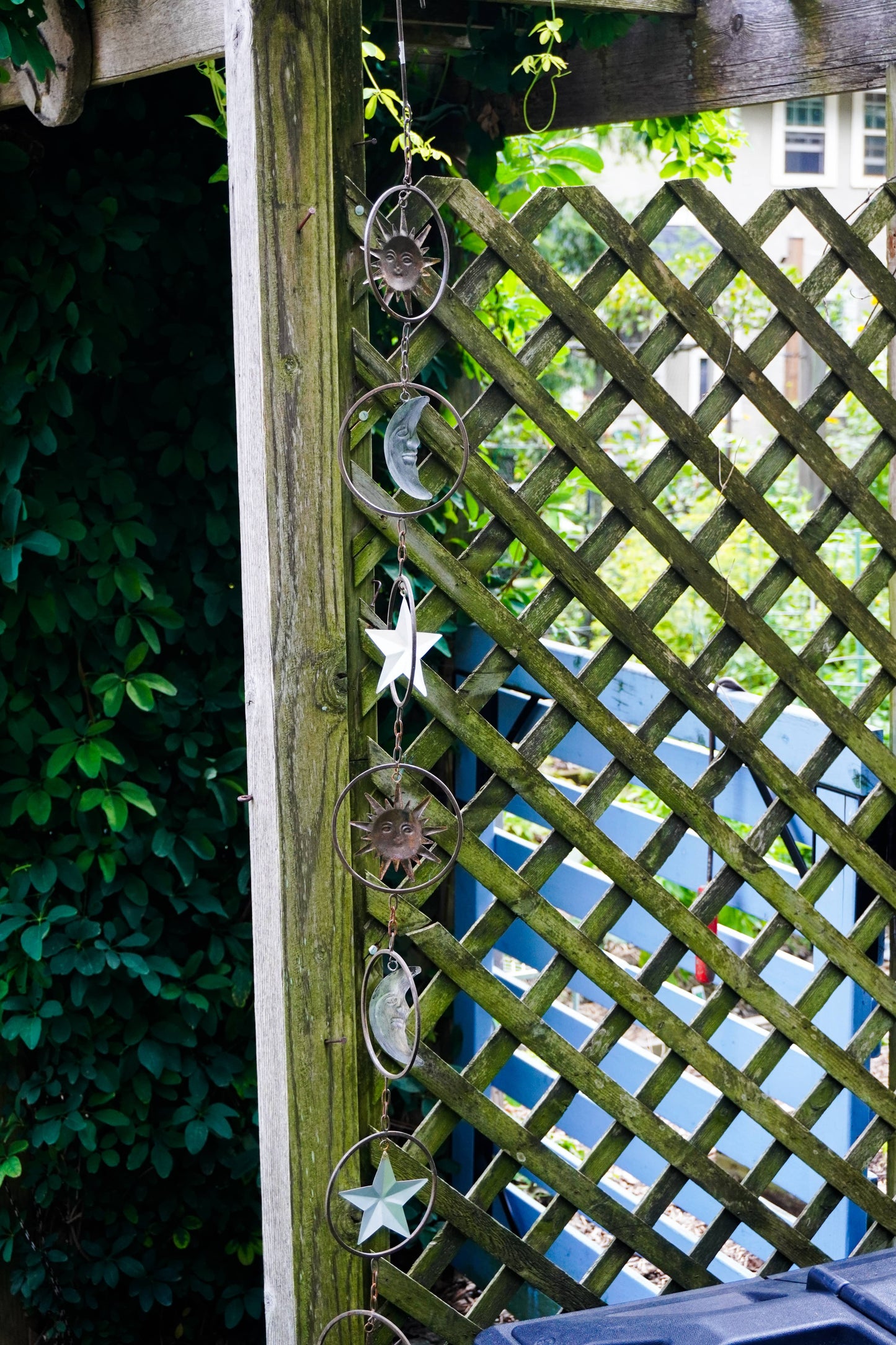 A vertical garden trellis post decorated with a hanging string of metal celestial ornaments, including stars, moons, and sunbursts, next to a wooden lattice fence and green foliage.
