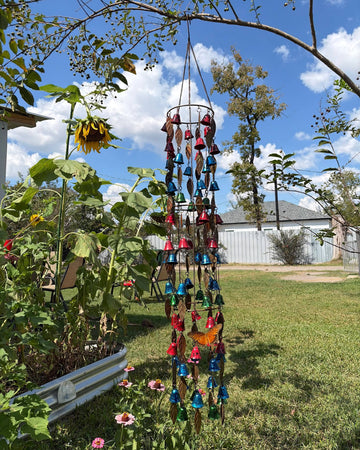 A colorful wind chime garden hanging suspended from a tree branch in a backyard garden. The wind chime features multiple tiers with blue, red, and gold bells, small wind chimes, and butterfly decorations.