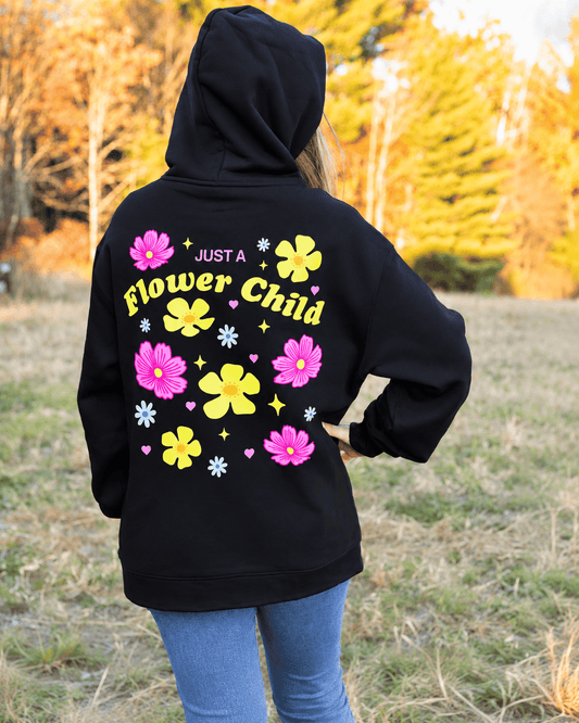 A person standing outdoors in a grassy field wearing a black hoodie from the back, which features a colorful graphic print of pink and yellow flowers and the text 'JUST A Flower Child' in yellow.