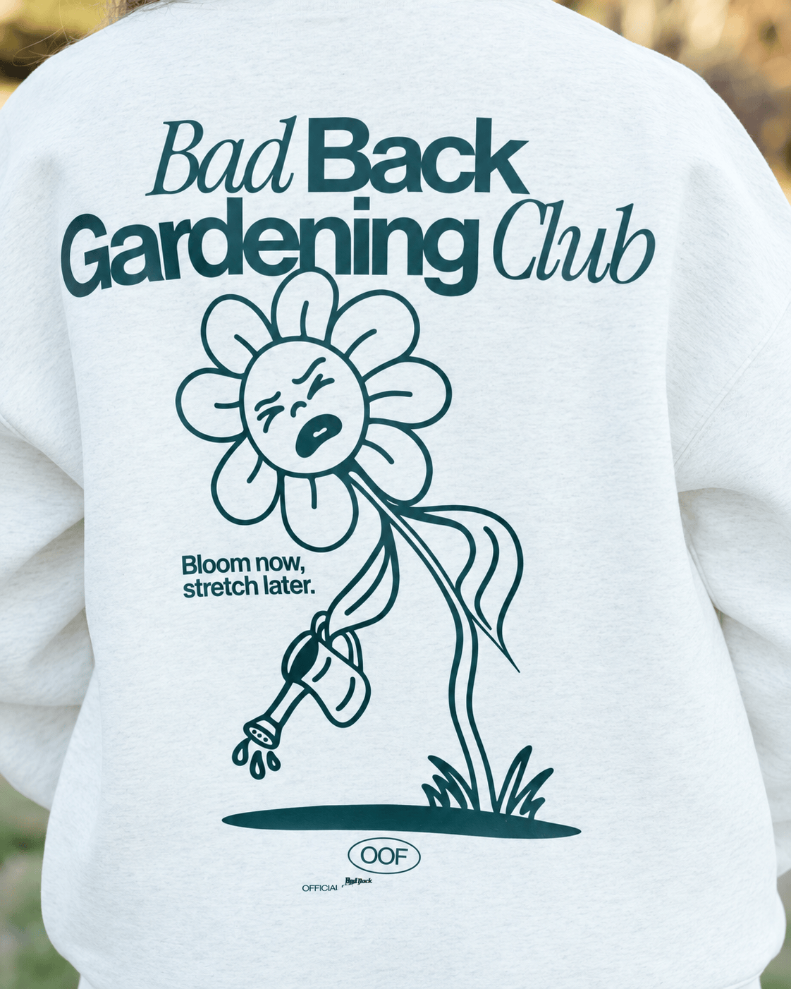 A close-up of the back of a white sweatshirt worn by a person, featuring a dark teal graphic of a sad, thirsty flower watering itself with a hose, with the text 'Bad Back Gardening Club' and 'Bloom now, stretch later.'.