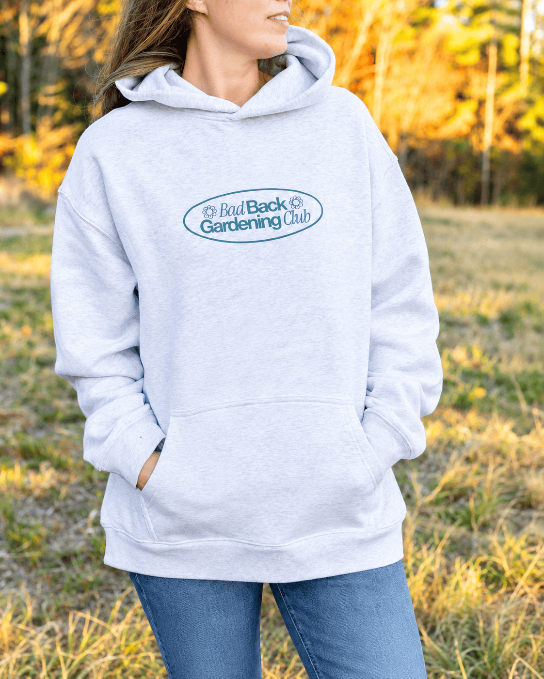 A woman wearing a light heather gray pullover hoodie with a blue-green oval 'Real Back Gardening Club' logo on the chest, standing outdoors in a grassy field.