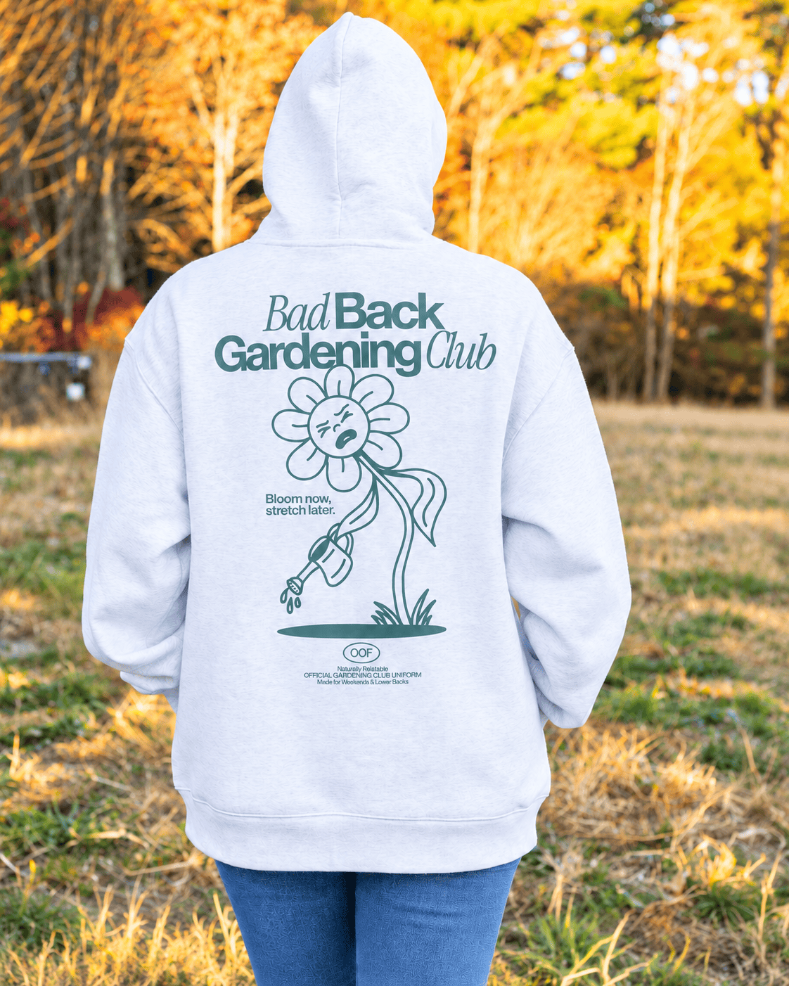 A person standing outdoors in a grassy field at sunset, viewed from behind, wearing a white hooded sweatshirt with a large green graphic on the back that reads 'Bad Back Gardening Club' and shows blue jeans.