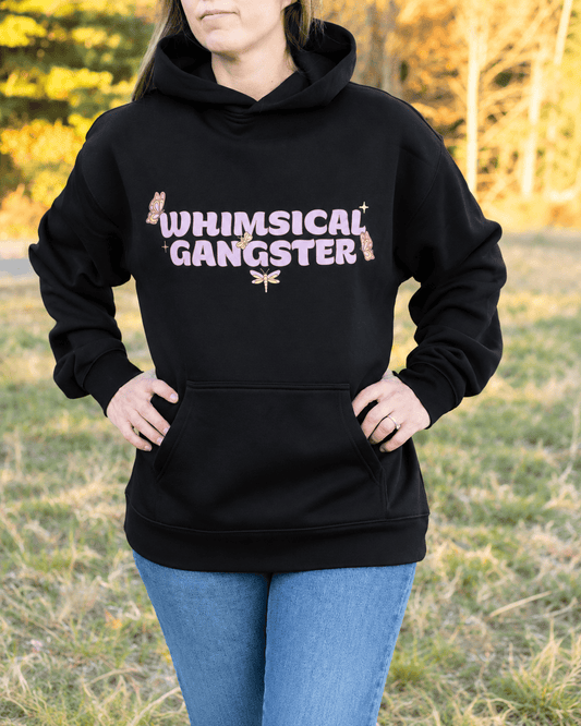 A woman wearing a black pullover hoodie with 'WHIMSICAL GANGSTER' printed in pink glitter-style letters with small butterfly and dragonfly graphics, paired with blue jeans, standing outdoors in a grassy field.
