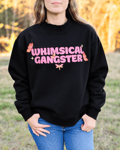 A woman wearing a black crewneck sweatshirt with a pink graphic that reads 'WHIMSICAL GANGSTER' decorated with pink gummy bears and a dragonfly, paired with blue jeans in an outdoor setting.