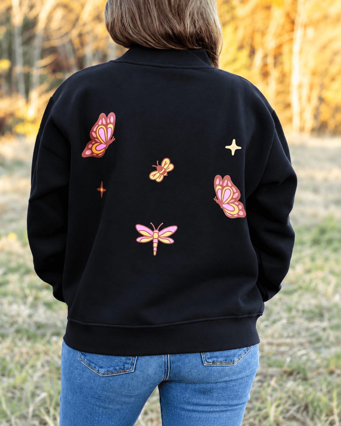 A person viewed from behind wearing a black crewneck sweatshirt with colorful embroidered butterflies and stars on the back, paired with blue jeans, set against a blurred outdoor background.