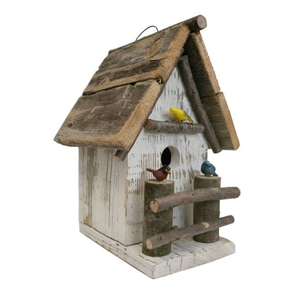 A rustic birdhouse made from weathered wood, featuring a white-washed body, a slanted wooden roof with a hanging loop, a small round entrance hole, a perch, and two log-style supports. Three small decorative birds are perched on the structure: one yellow, one reddish-brown, and one blue-gray.