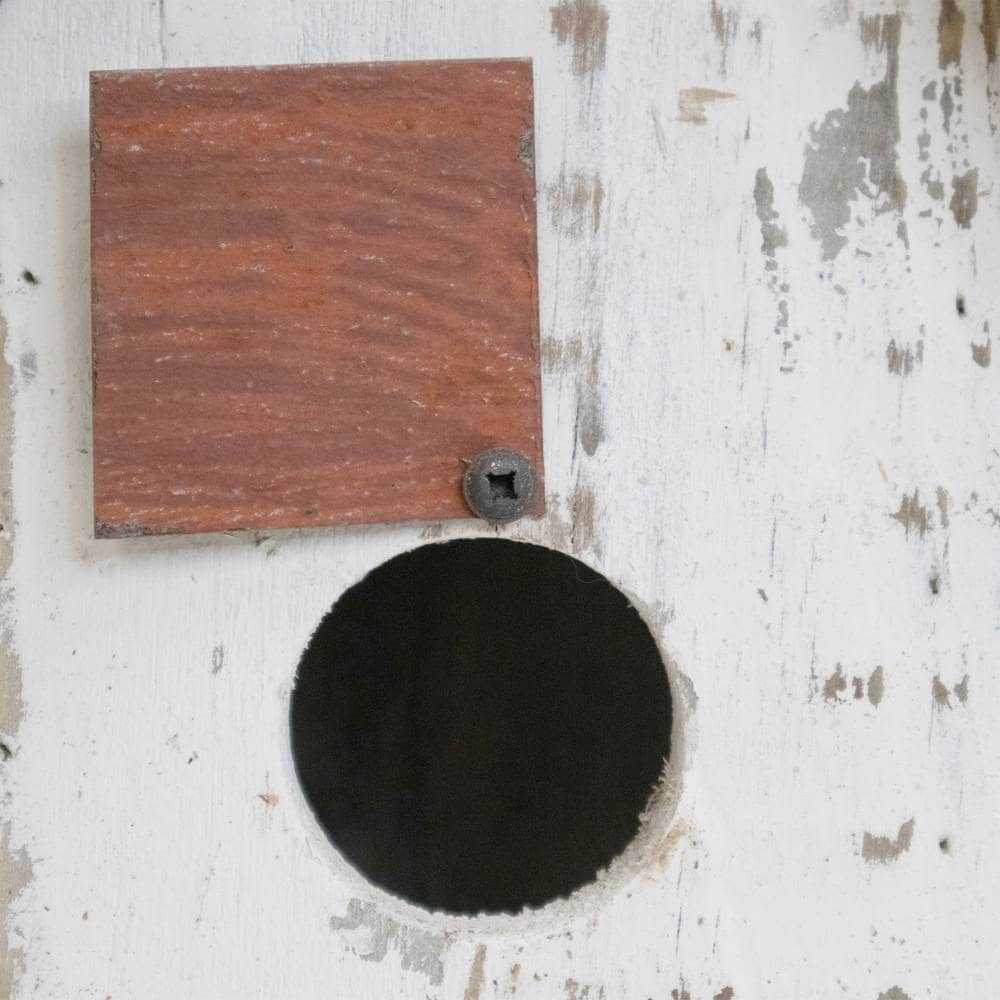 A square reddish-brown wooden board with a single screw, mounted on a distressed white painted wooden surface next to a circular black opening.