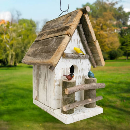 A rustic white-painted wooden birdhouse with a weathered brown roof and perch, hanging outdoors in a grassy park. Three small decorative birds are perched on the birdhouse.