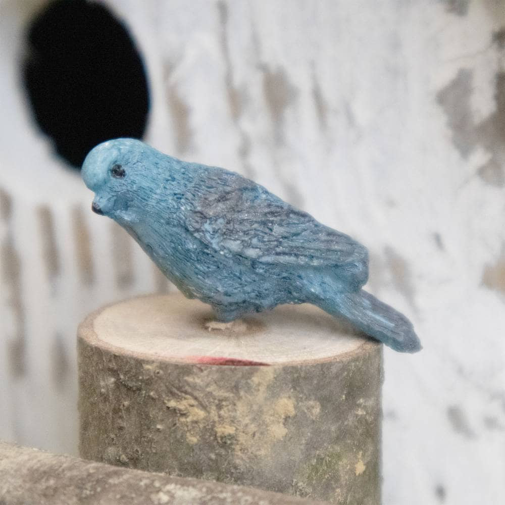 A small blue bird figurine with a textured, stone-like finish, perched on a rustic wooden log against a white stucco wall with a circular hole.