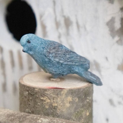 A small blue bird figurine with a textured, stone-like finish, perched on a rustic wooden log against a white stucco wall with a circular hole.