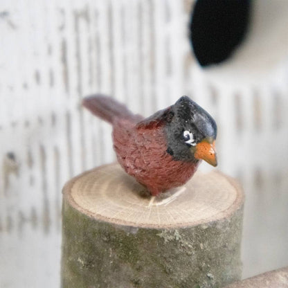 A small bird figurine with a black head, reddish-brown body, and orange beak perched on a cut wood stump against a soft white background.