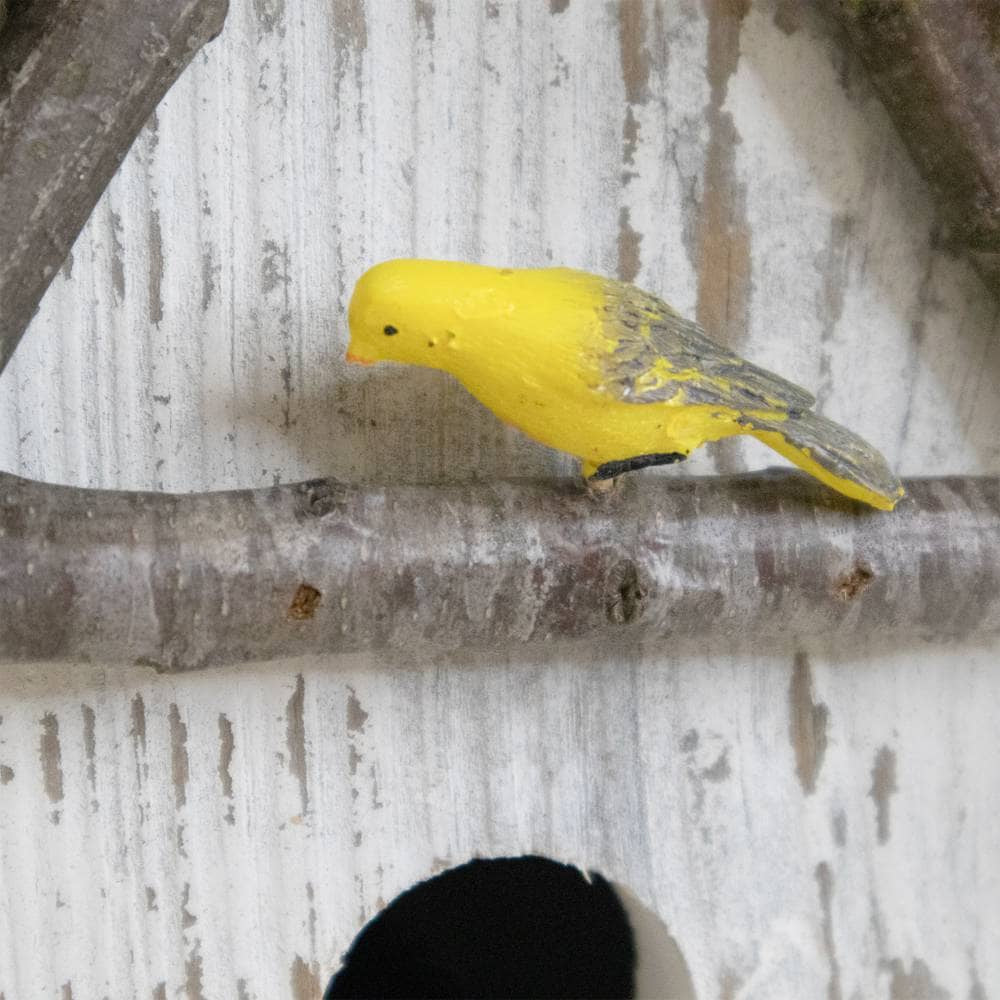 A small yellow bird figurine with gray wings perched on a rustic tree branch in front of a distressed white wooden background.