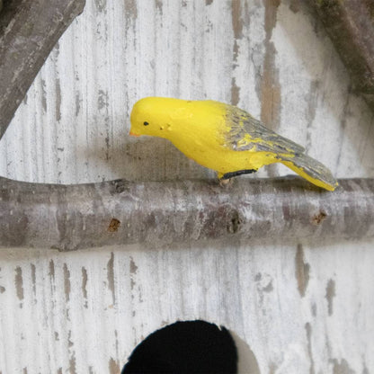A small yellow bird figurine with gray wings perched on a rustic tree branch in front of a distressed white wooden background.