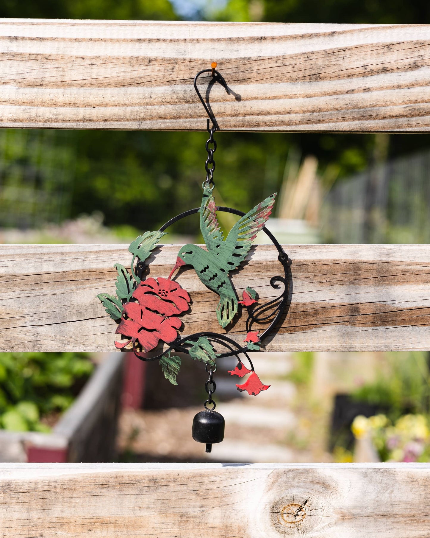 Metal hummingbird wind chime ornament with red flowers and green leaves, hanging from a wooden fence by a black S-hook