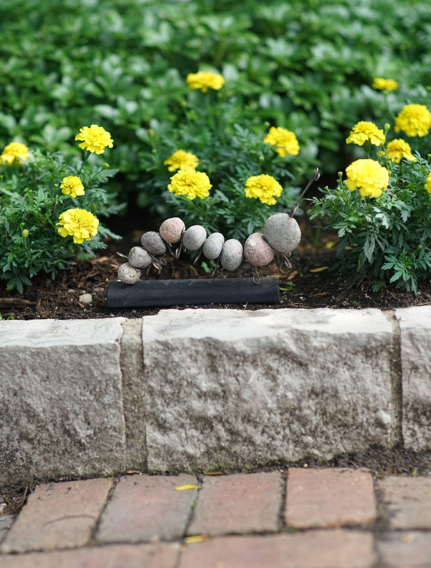 Stone pebble caterpillar garden sculpture on a black base displayed in a flower bed with yellow marigolds