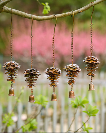Five copper metal pine cone wind chimes with small brass bells hanging from a tree branch outdoors