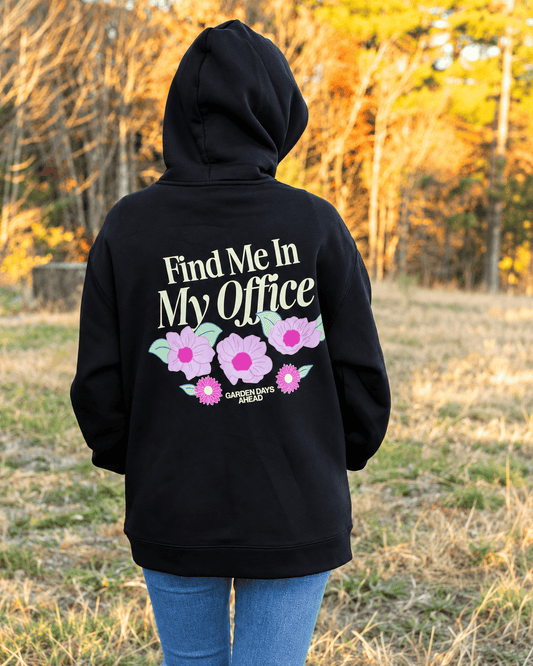 A person viewed from behind wearing a black hoodie with a graphic on the back that reads 'Find Me In My Office' above pink flowers and the words 'Garden Days Ahead,' standing outdoors in a grassy area with autumn trees in the background.