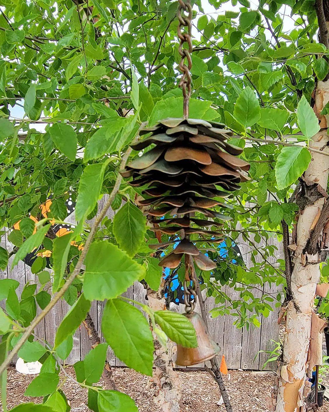 A metal pine cone wind chime hanging from a tree branch in a backyard. The feeder features a stacked, tiered design resembling pine cones in a weathered bronze-brown finish, with a small hanging metal bell below.