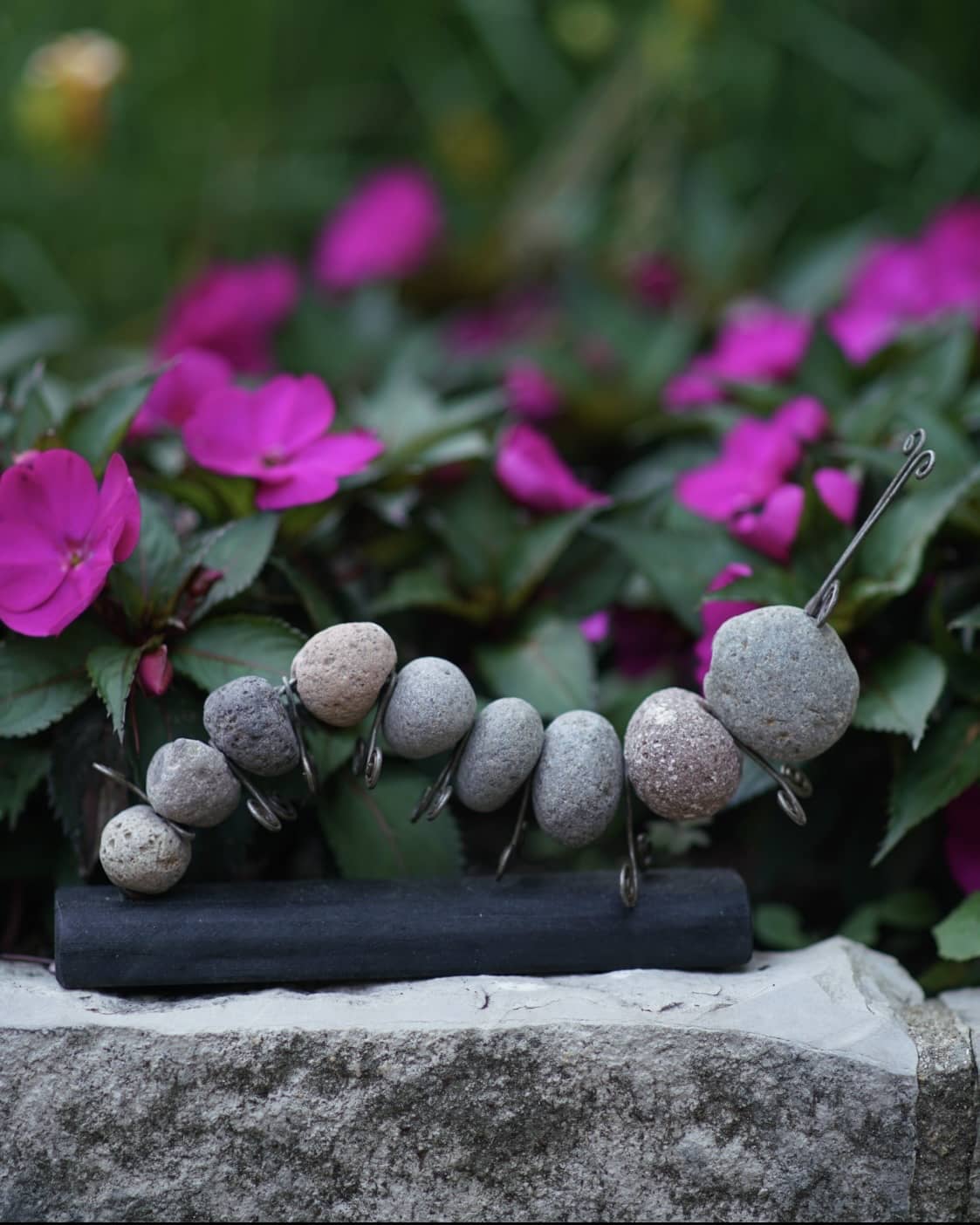 Rock and wire caterpillar garden sculpture on a dark wooden base, displayed outdoors near pink flowers