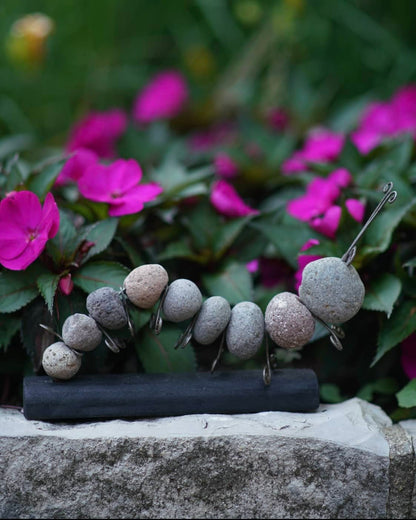 Rock and wire caterpillar garden sculpture on a dark wooden base, displayed outdoors near pink flowers