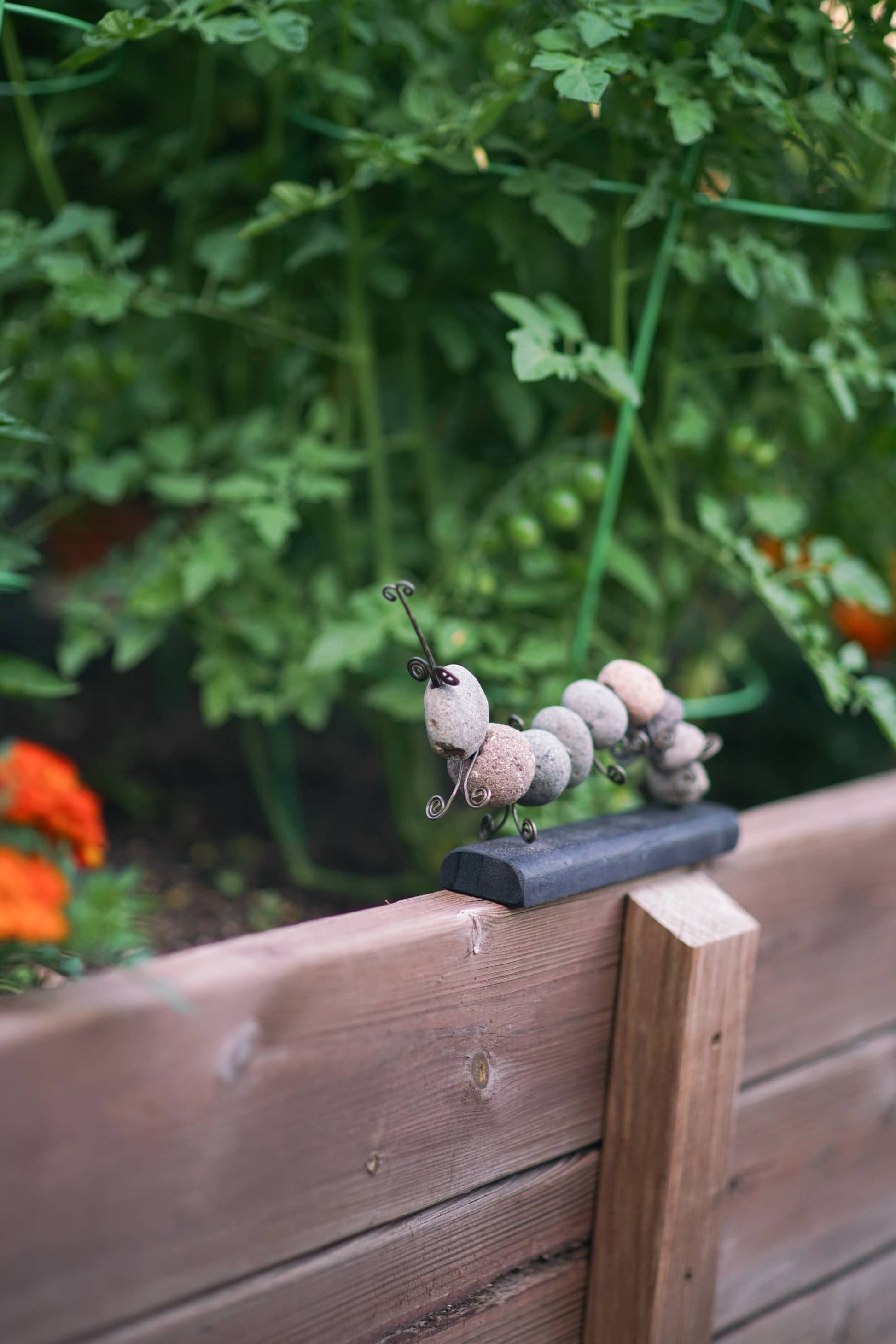 Stone caterpillar sculpture with wire antennae and legs on a dark base, perched on a wooden raised garden bed