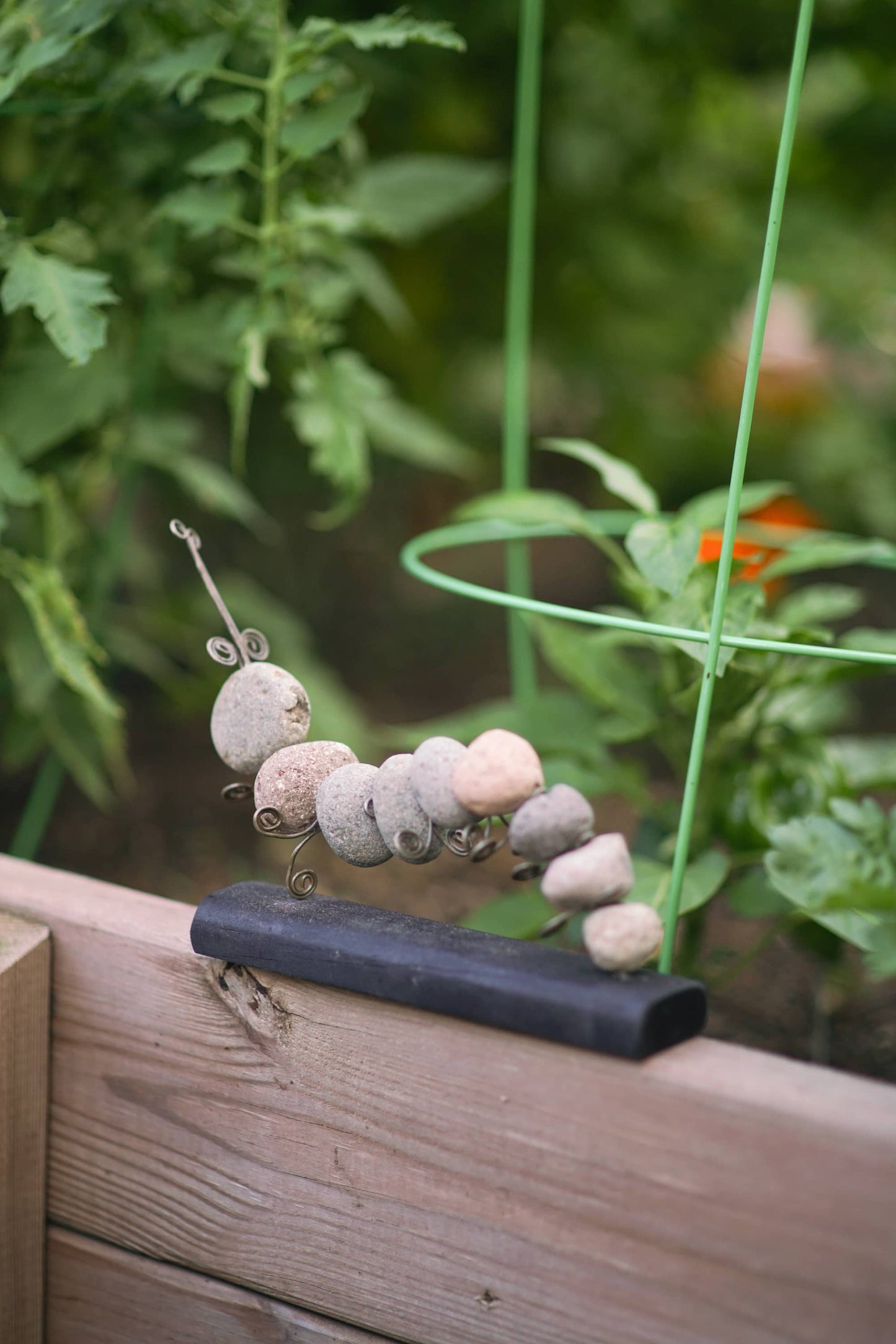 Rock and wire caterpillar garden sculpture with spiral antennae mounted on a dark rectangular base, displayed on a wooden raised garden bed