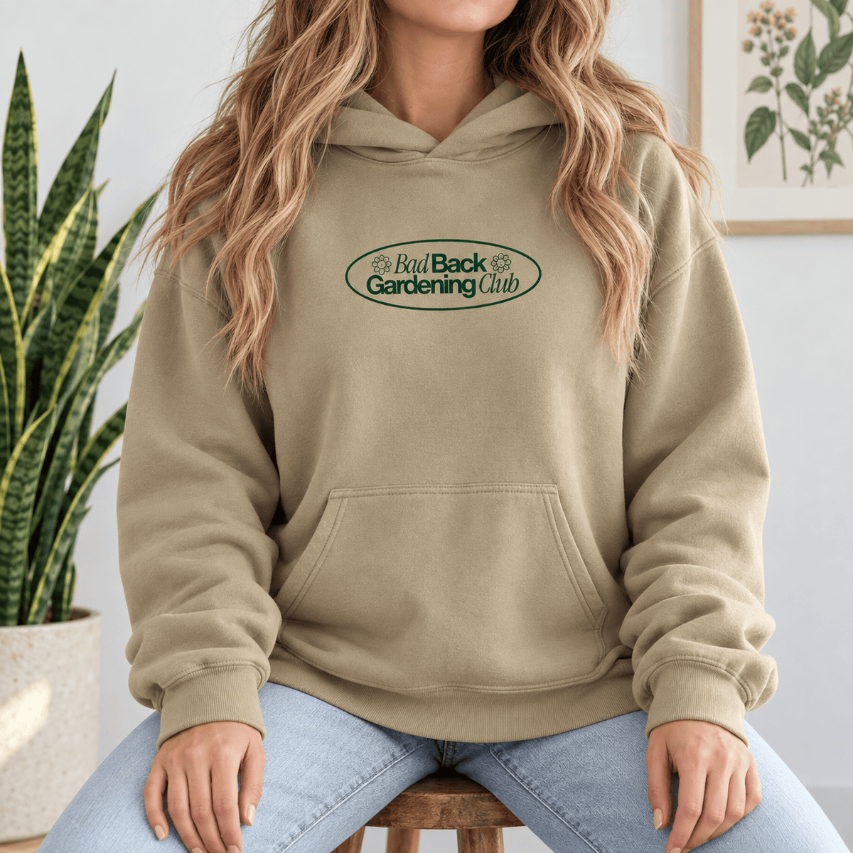 A woman wearing a beige pullover hoodie with a green and white 'Bad Back Gardening Club' logo on the chest, sitting on a wooden stool in a bright room.