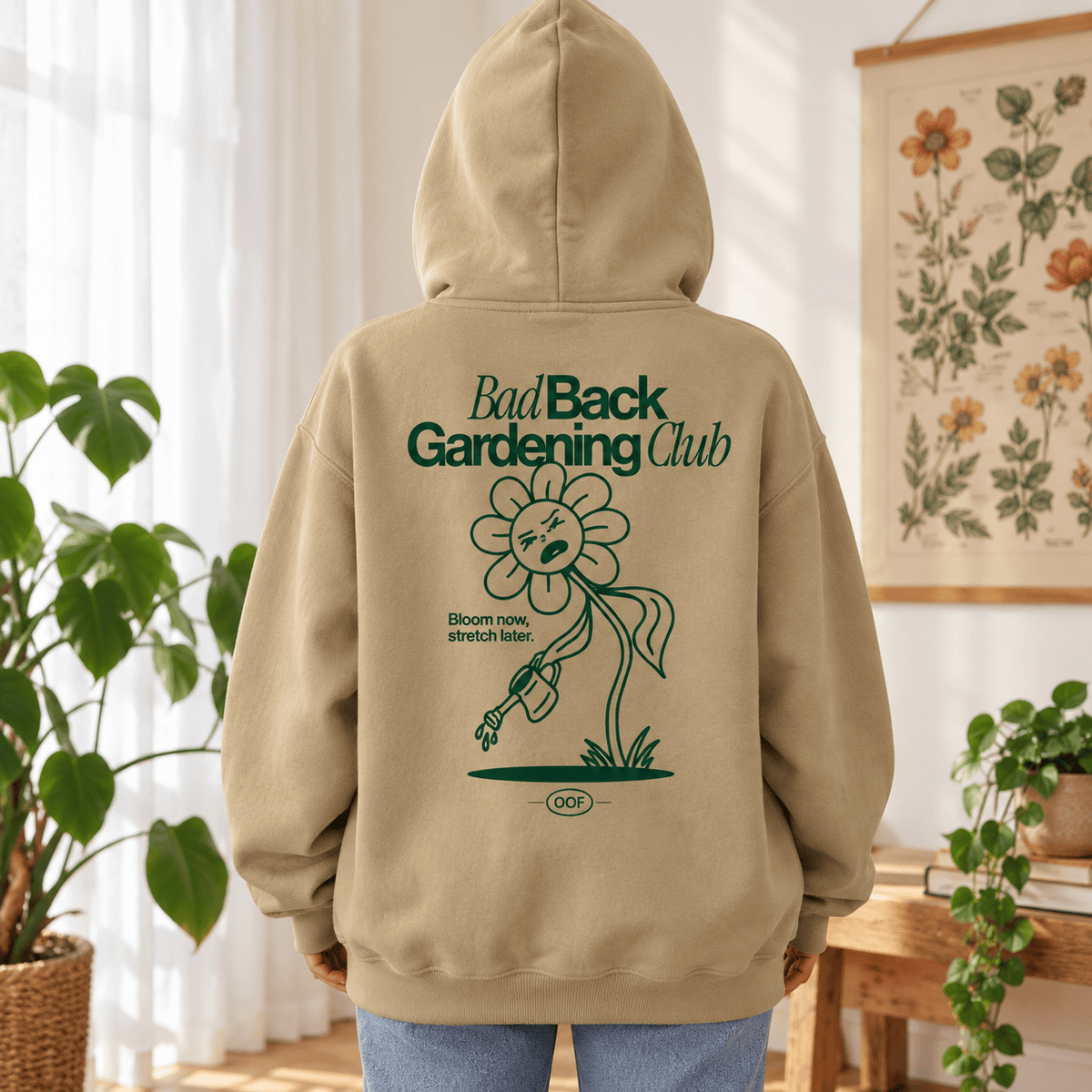 A person shown from the back wearing a beige hoodie with a green graphic print that reads 'Bad Back Gardening Club' and features a flower illustration. The setting is a bright, sunlit room with plants and a floral wall hanging.