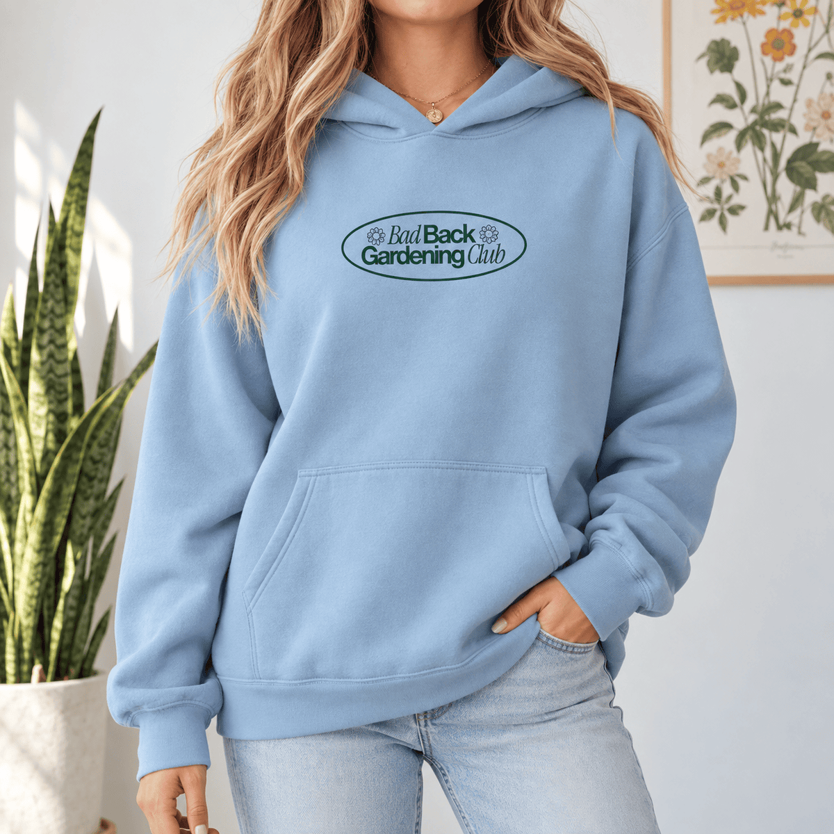 A woman wearing a light blue hoodie with a 'Bad Back Gardening Club' graphic on the chest, paired with light-wash jeans in a bright indoor setting.