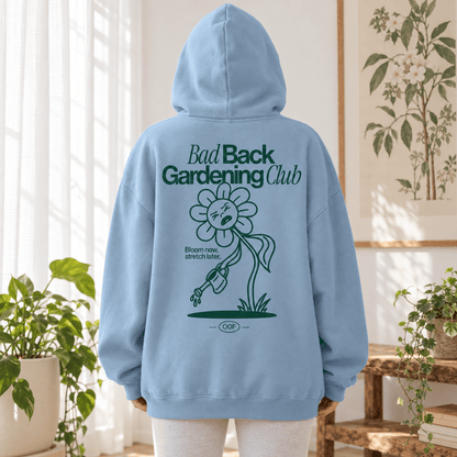A person wearing a light blue hoodie from the back, standing in a bright indoor room with plants and a framed botanical print. The hoodie has a large dark green graphic on the back reading 'Bad Back Gardening Club' with a flower illustration and small text below.