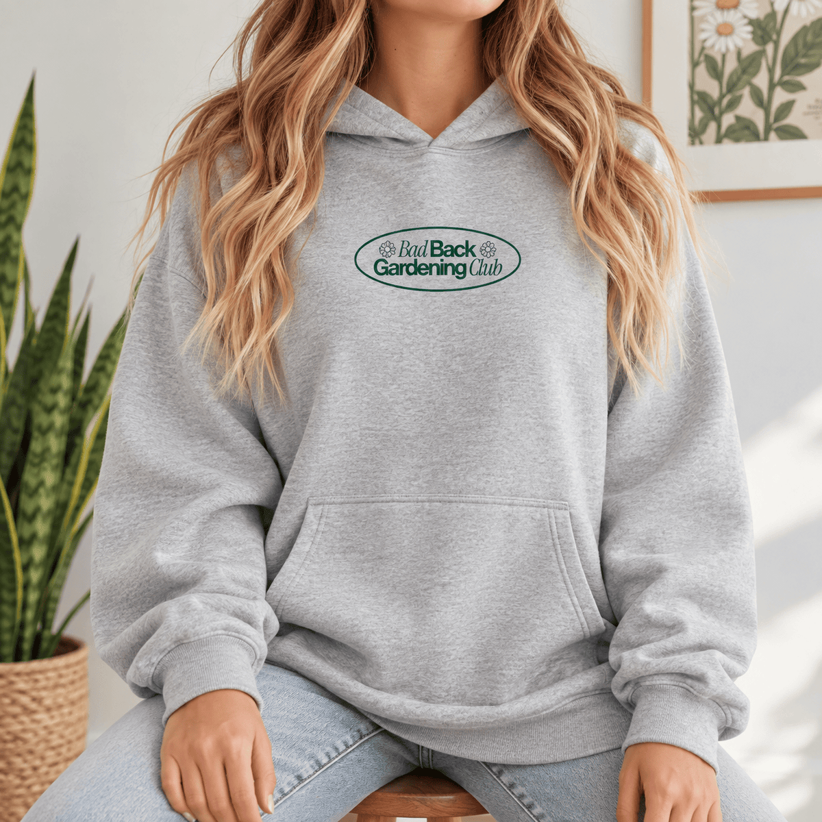 A woman wearing a light gray pullover hoodie with an oval 'Bad Back Gardening Club' logo on the chest, paired with light-wash jeans in a bright indoor setting.