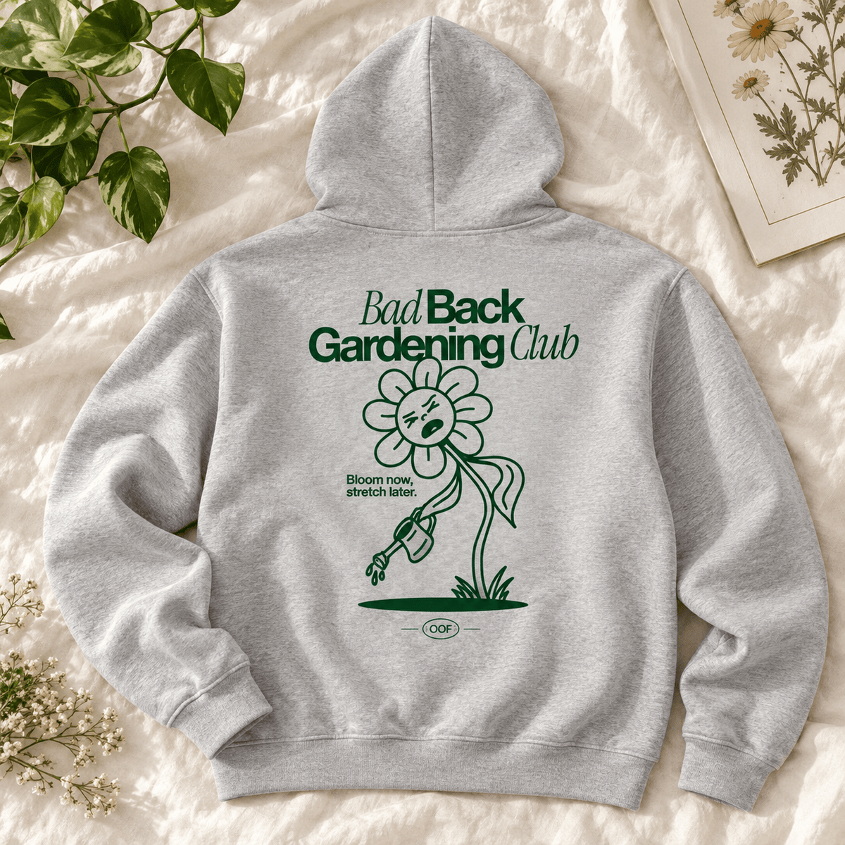 A light gray heather hoodie laid flat on a white textured fabric background, surrounded by decorative botanical props. The back of the hoodie features a dark green graphic of a flower with a sad face and the text 'Bad Back Gardening Club' and 'Bloom now, stretch later.
