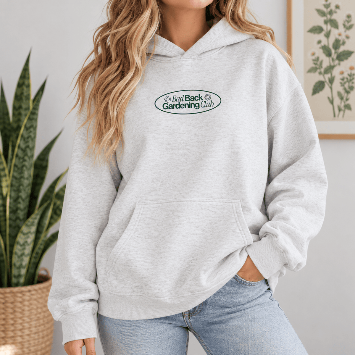 A woman wearing a light heather gray hoodie with a green and black oval chest graphic that reads 'Bad Back Gardening Club'. She is also wearing light blue jeans, standing in an indoor setting with a snake plant on the left and a framed botanical print on the right.