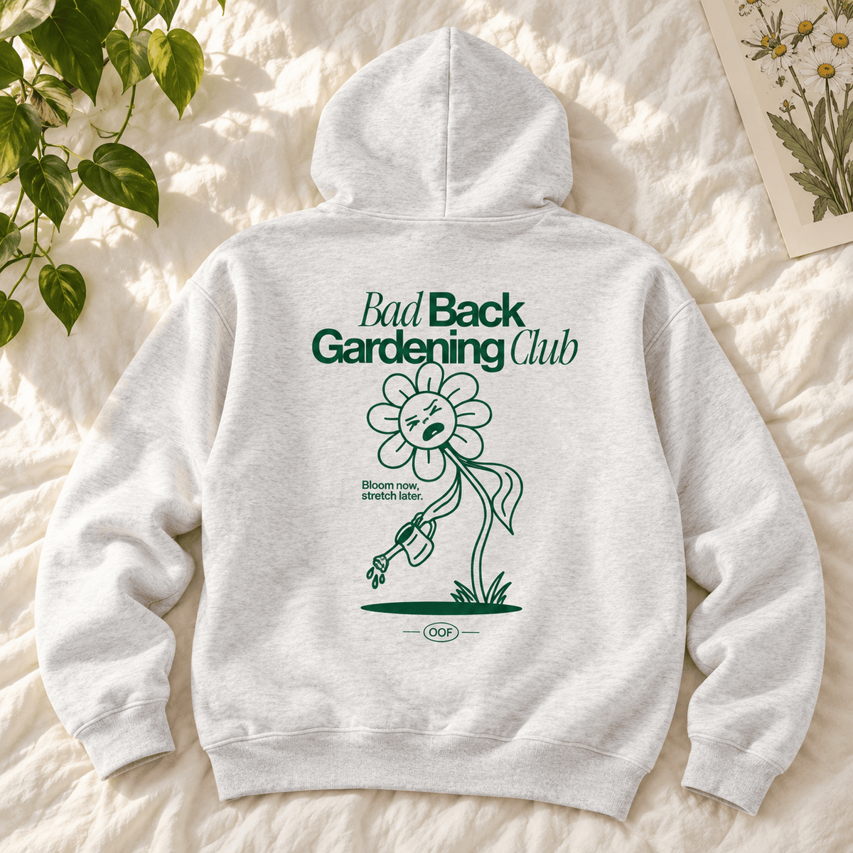 A light heather gray hoodie laid flat on a textured cream fabric background, with a green graphic on the back reading 'Bad Back Gardening Club' and a stylized flower illustration. Green leaves and part of a botanical card are visible around the edges.