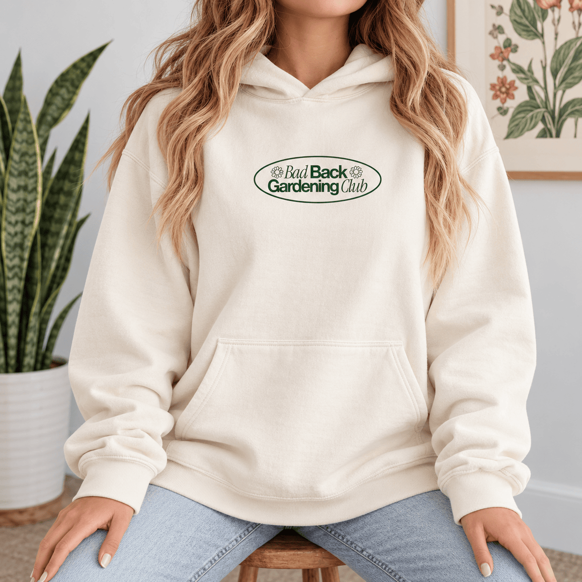 A woman wearing a cream-colored pullover hoodie with a green and black 'Bad Back Gardening Club' logo on the chest, paired with light blue jeans, sitting in a home setting.
