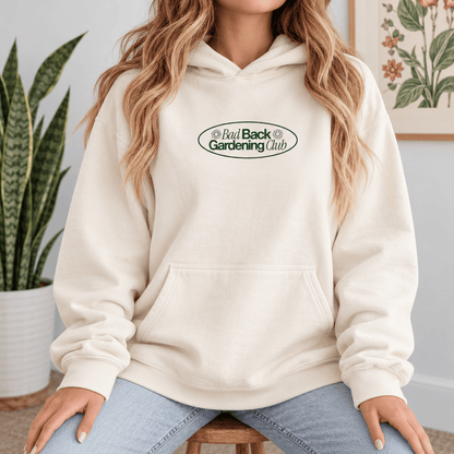A woman wearing a cream-colored pullover hoodie with a green and black 'Bad Back Gardening Club' logo on the chest, paired with light blue jeans, sitting in a home setting.