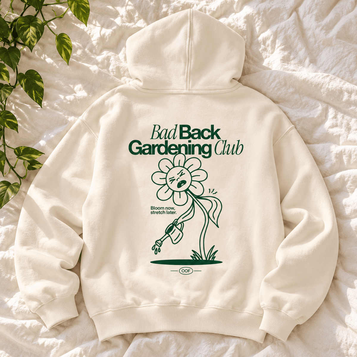 An off-white hooded sweatshirt laid flat on a textured white fabric background, with green printed text reading 'Bad Back Gardening Club' and a cartoon flower graphic on the back. A leafy plant is visible at the top left.