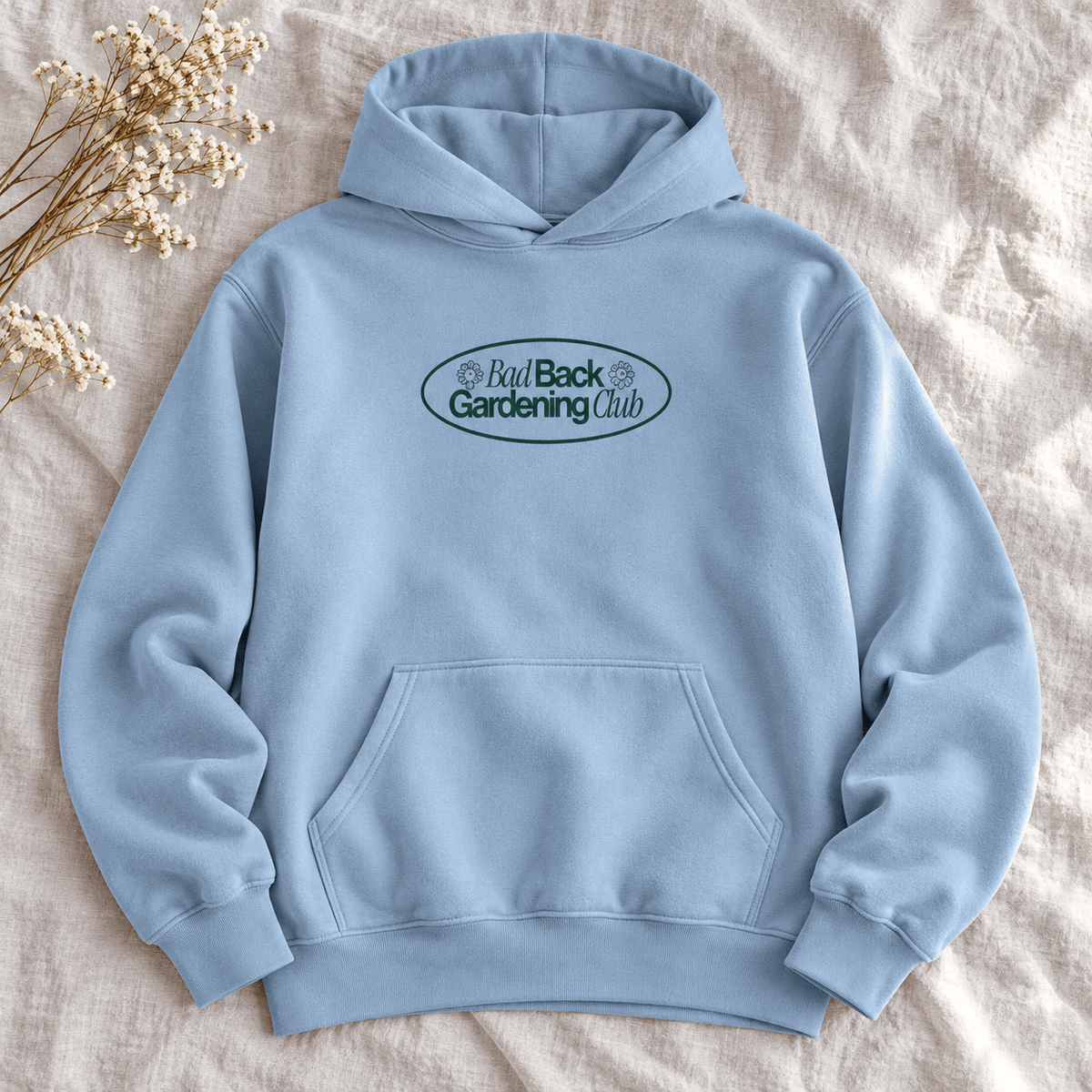 A light blue pullover hoodie laid flat on a textured beige fabric background, with a small chest graphic reading 'Bad Back Gardening Club' inside an oval. A small dried floral arrangement sits in the upper left corner.