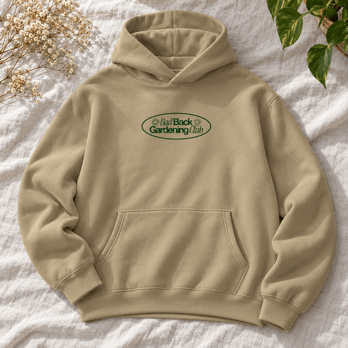 A beige pullover hoodie with a front kangaroo pocket and a green oval chest graphic reading 'Bad Back Gardening Club,' styled flat on a white textured fabric with dried flowers and green leaves in the corners.