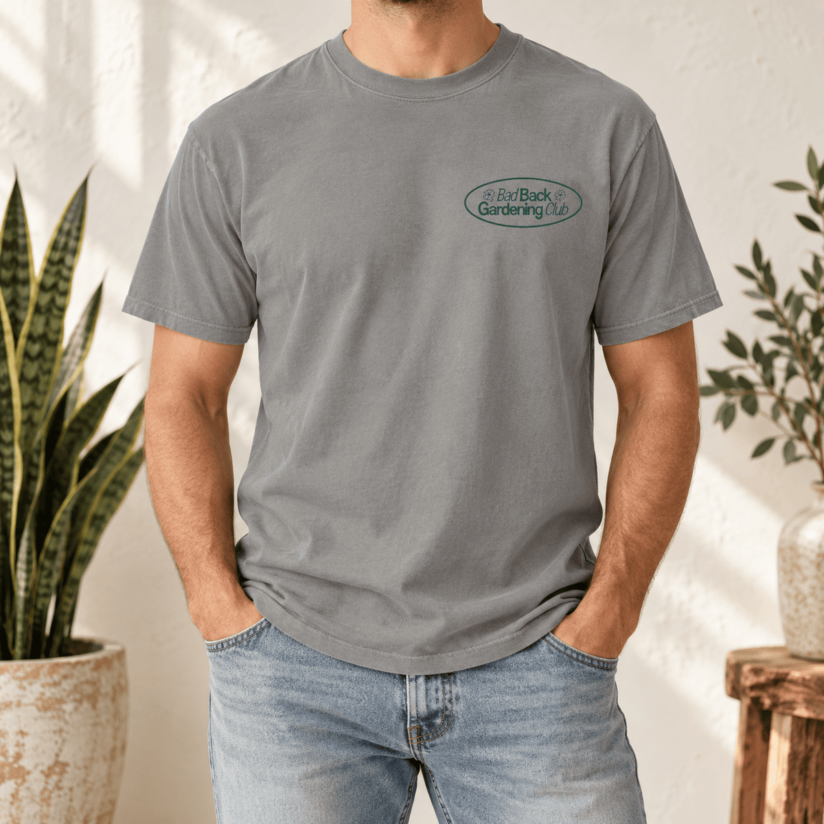 A man wearing a heather gray crewneck t-shirt with a small green and white logo on the left chest, paired with light blue jeans in a bright, sunlit indoor setting with potted plants.