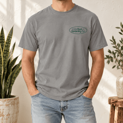 A man wearing a heather gray crewneck t-shirt with a small green and white logo on the left chest, paired with light blue jeans in a bright, sunlit indoor setting with potted plants.