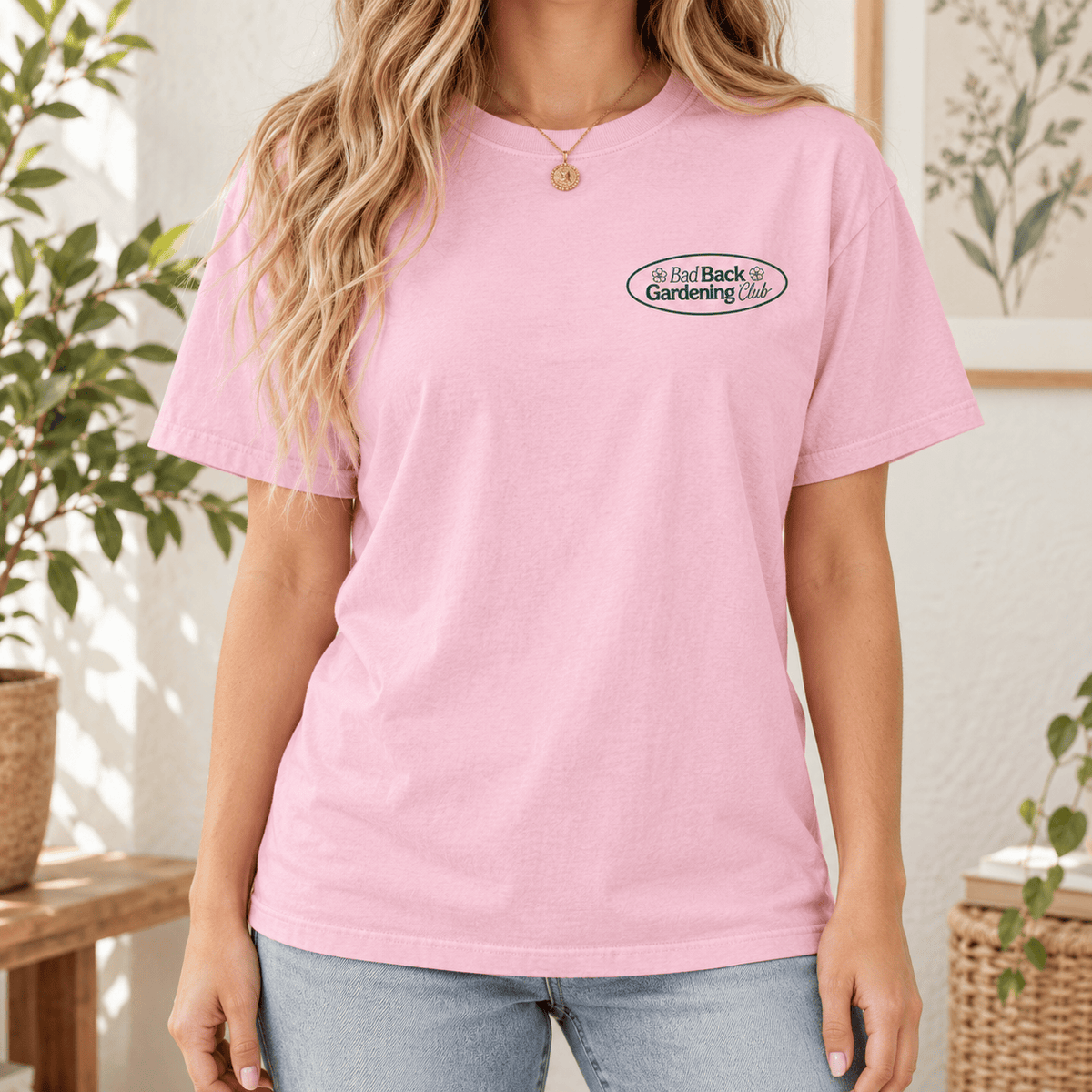 A woman wearing a light pink crewneck t-shirt with a small 'Bad Back Gardening' logo on the left chest, paired with light blue jeans in a bright, sunlit indoor setting.