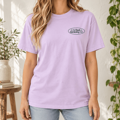 A woman wearing a light lavender crewneck t-shirt with a small oval 'Bad Back Gardening Club' logo on the left chest, paired with light blue jeans in a sunlit indoor setting with plants.