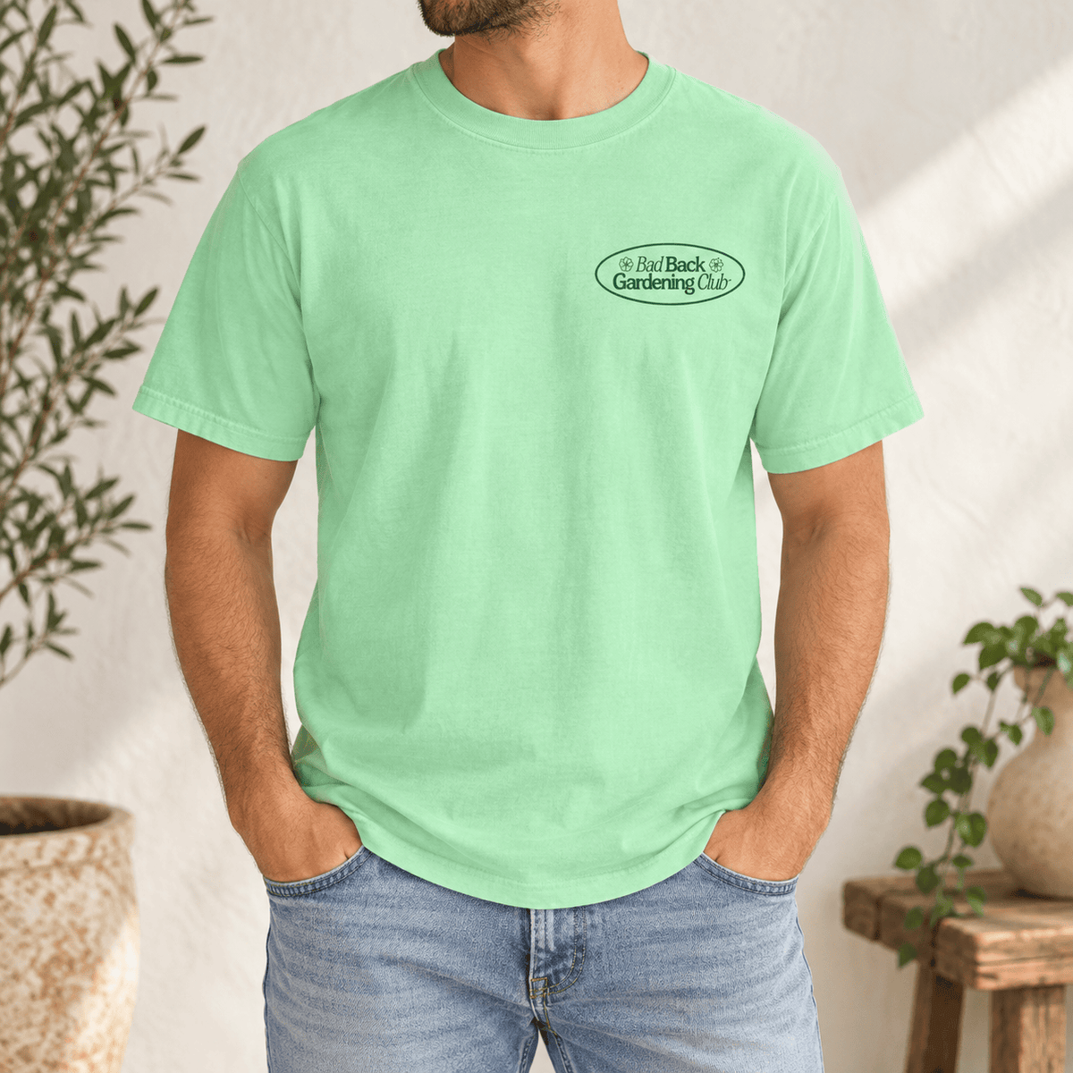 A man wearing a mint green crewneck t-shirt with a small oval 'Bad Back Gardening Club' logo on the left chest, paired with light blue jeans, set against a warm, sunlit wall with potted plants.