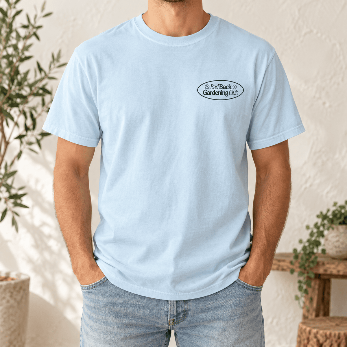 A man wearing a light blue crewneck t-shirt with a small black oval logo on the left chest that reads 'Bad Back Gardening Club'. He is also wearing light-wash blue jeans, standing in a brightly lit room with a textured wall and potted plants.